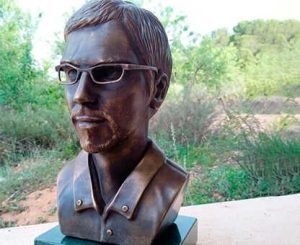 A bronze bust of a man wearing glasses and a collared shirt, displayed outdoors with greenery in the background.