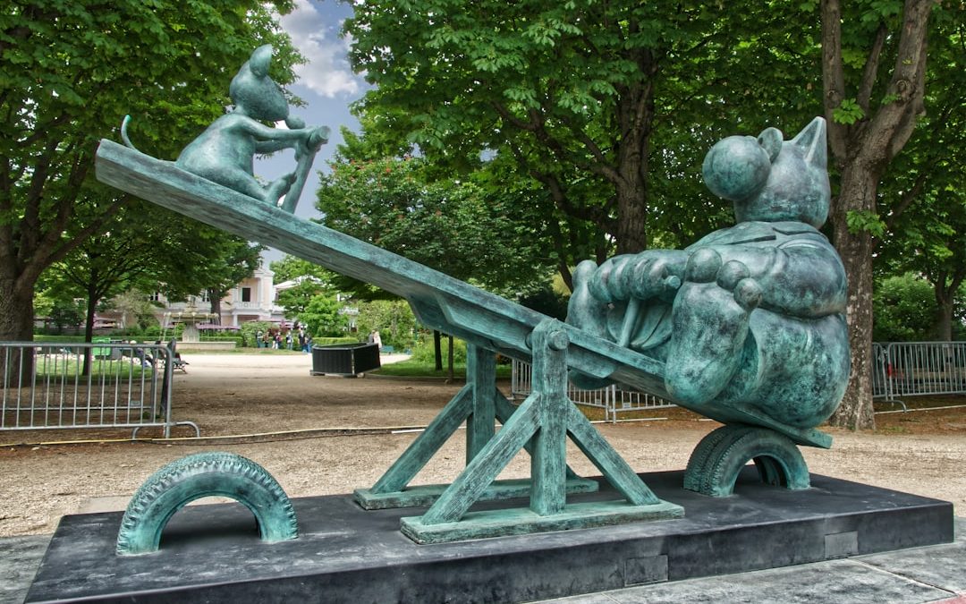 Bronze statue in a park depicting a cat and a kitten on a seesaw, with the larger cat sitting and the kitten standing. Trees and a fenced area are visible in the background.