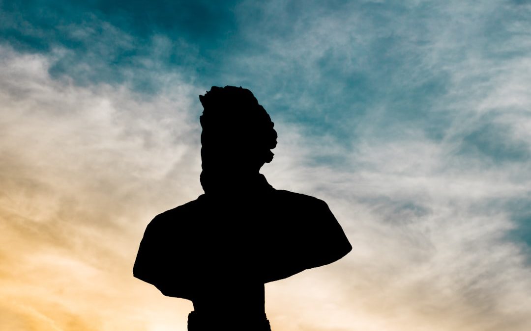 Silhouette of a classical statue bust against a sky with mixed clouds and colors at sunrise or sunset.